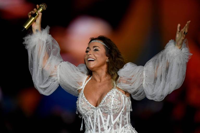 Uma mulher está se apresentando em um palco, vestindo um vestido branco com mangas bufantes e detalhes brilhantes. Ela segura um microfone dourado e sorri, com os braços levantados em um gesto de celebração. O fundo é desfocado, mas sugere um ambiente de show com luzes e uma multidão.