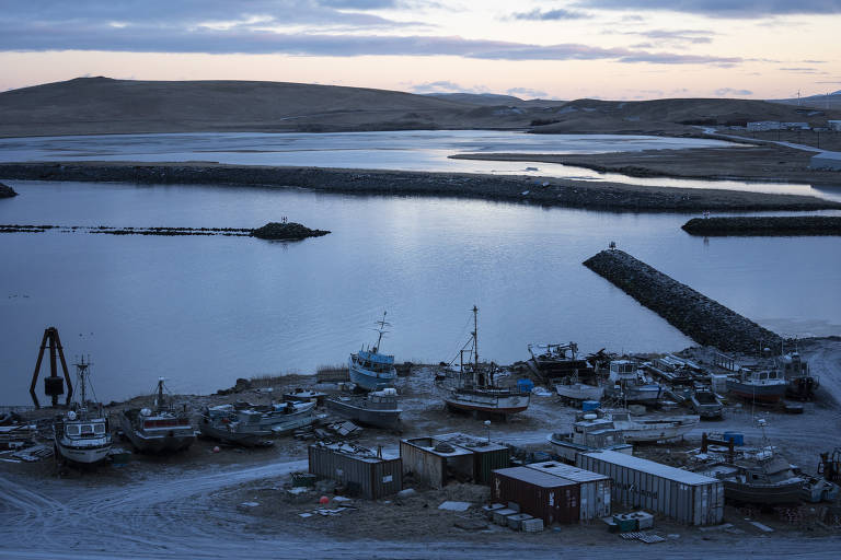 A imagem mostra um porto com vários barcos de pesca encalhados em uma área de areia. O cenário é tranquilo, com água calma refletindo o céu nublado. Ao fundo, há uma pequena elevação de terra e um quebra-mar que se estende na água. O ambiente parece ser rural e isolado, com poucos sinais de atividade humana.