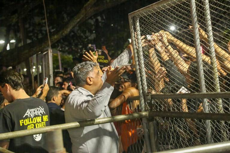 A imagem mostra um grupo de pessoas em um evento musical, com algumas delas estendendo as mãos através de uma grade de proteção. Um homem de camisa clara está se comunicando com o público, enquanto outros na multidão seguram celulares. Ao fundo, há pessoas com camisetas escuras, algumas com mensagens visíveis. A atmosfera parece ser de entusiasmo e interação entre os artistas e os fãs.