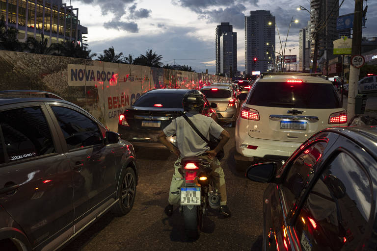A imagem mostra um congestionamento em uma avenida, com vários carros parados. Um motociclista está posicionado entre os veículos, olhando para frente. Ao fundo, há prédios altos e um céu nublado, indicando que é ao entardecer. Um painel publicitário é visível na lateral da estrada, e as luzes dos carros estão acesas, refletindo um ambiente urbano.