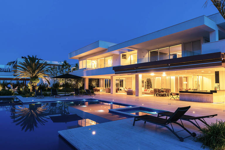 A imagem mostra uma casa moderna iluminada à noite, com uma piscina em primeiro plano. A estrutura da casa é de dois andares, com grandes janelas que refletem a luz interna. Há uma área externa com mesas e cadeiras, além de plantas ao redor. O céu está escuro, indicando que é noite.