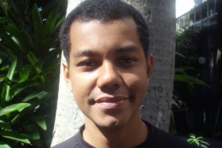 Um jovem homem está posando para a foto em um ambiente ao ar livre. Ele tem cabelo curto e liso, e está usando uma camiseta preta. Ao fundo, há uma árvore e folhagens verdes, indicando um ambiente natural e ensolarado.