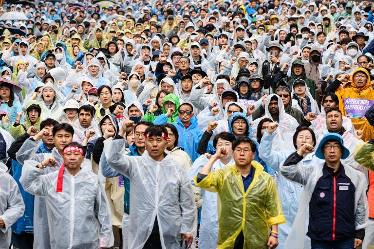 A imagem mostra uma grande multidão de pessoas reunidas em um evento ao ar livre, todas vestindo capas de chuva. Muitas delas estão levantando o punho em sinal de protesto ou apoio. O ambiente parece ser de uma manifestação, com uma variedade de cores nas capas de chuva, que vão do transparente ao colorido. O público é diversificado, com homens e mulheres de diferentes idades.