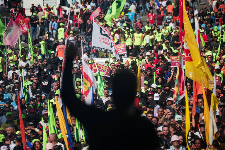A imagem mostra uma grande multidão reunida em uma manifestação. Muitas pessoas estão segurando bandeiras coloridas e cartazes, com destaque para camisetas de cor verde-limão. A cena é vista de um ângulo onde uma silhueta de uma pessoa levantando o braço é visível em primeiro plano, enquanto a multidão se estende ao fundo, repleta de pessoas e bandeiras.