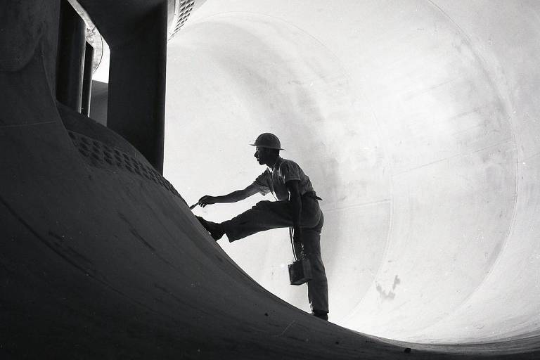 A imagem em preto e branco mostra um trabalhador dentro de um tubo de concreto. Ele está em uma posição de equilíbrio, com uma perna levantada e a outra apoiada no tubo. O trabalhador usa um capacete e segura uma ferramenta na mão, enquanto parece estar realizando algum tipo de trabalho na superfície interna do tubo. O ambiente é amplo e as paredes do tubo são lisas e arredondadas, criando um efeito visual interessante com a luz que entra.