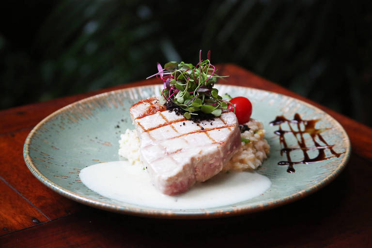 Um prato de cerâmica com um pedaço de peixe grelhado, decorado com ervas e um tomate-cereja. O peixe está sobre uma cama de arroz e acompanhado por um molho branco e um fio de redução de balsâmico. O fundo é desfocado, sugerindo um ambiente natural.