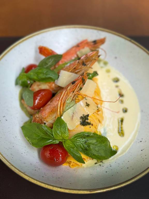 Um prato gourmet apresentado em uma tigela branca. O prato contém camarões grelhados, tomates cereja, folhas de manjericão e lascas de queijo. Há um molho cremoso ao redor, com um toque de azeite e ervas.