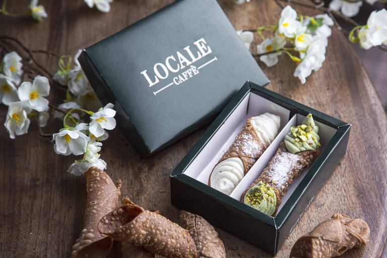 A imagem mostra uma caixa verde com a inscrição 'LOCALE CAFFÈ' na parte superior. Dentro da caixa, há dois canolis, um com recheio branco e outro com recheio verde, ambos polvilhados com açúcar. Ao redor da caixa, há canolis quebrados e flores brancas decorativas.