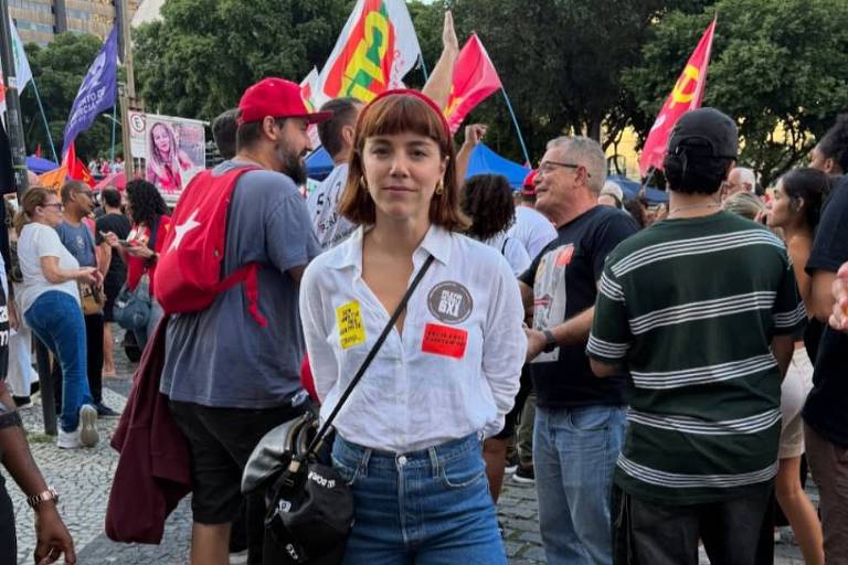 A imagem mostra uma jovem mulher em um ambiente de manifestação. Ela está vestindo uma camisa branca e jeans, com um lenço vermelho na cabeça. Ao fundo, há um grupo de pessoas segurando bandeiras e cartazes, em um espaço urbano com prédios visíveis. O chão é de pedras e a atmosfera parece ser de protesto ou celebração.
