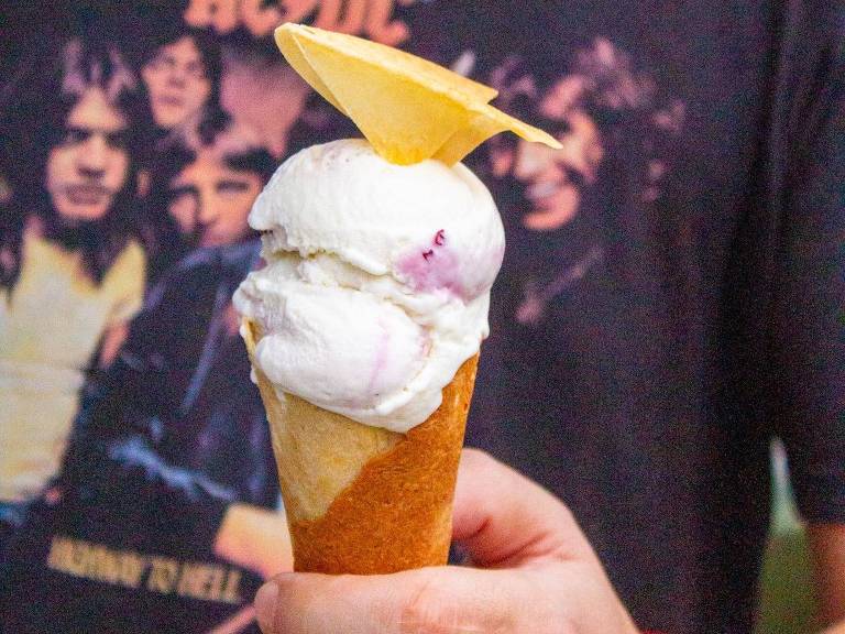 A imagem mostra uma pessoa segurando um cone de sorvete. O sorvete é de cor clara, com uma fatia de abacaxi em cima. A pessoa está usando uma camiseta preta com a estampa da banda AC/DC.