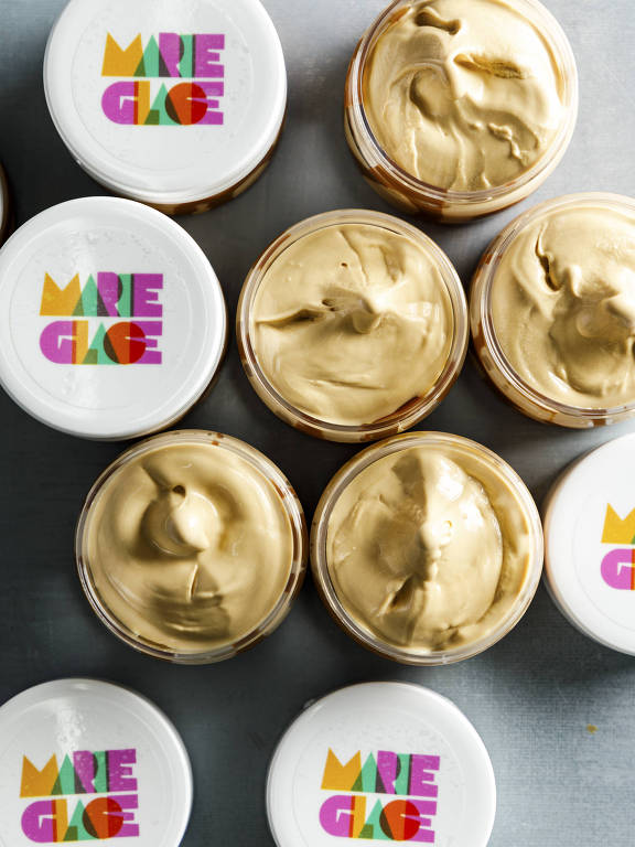 A imagem mostra vários potes de creme dispostos em uma superfície. Os potes têm tampas brancas com o texto colorido 'MARIE GLACE' em letras grandes e estilizadas. O conteúdo dos potes é de uma cor dourada cremosa, visível através das tampas abertas. A disposição dos potes é variada, com alguns potes em cima e outros embaixo, criando um padrão visual interessante.