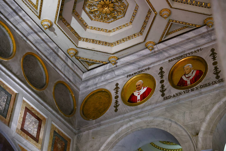 A imagem mostra o teto e as paredes de um ambiente com detalhes arquitetônicos elaborados. O teto é octogonal, decorado com um padrão dourado e um grande ornamento central. Nas paredes, há círculos com retratos de dois papas, um identificado como Francisco e o outro como Bento XVI, ambos em trajes papais. Abaixo dos retratos, há uma inscrição em latim que parece mencionar a duração do papado.