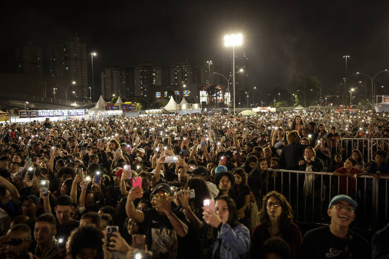 A imagem mostra uma grande multidão reunida em um evento noturno, com muitas pessoas segurando celulares com as luzes acesas. O ambiente é iluminado por refletores e há tendas ao fundo. A atmosfera é de animação e expectativa, com várias pessoas visíveis na frente e ao fundo.