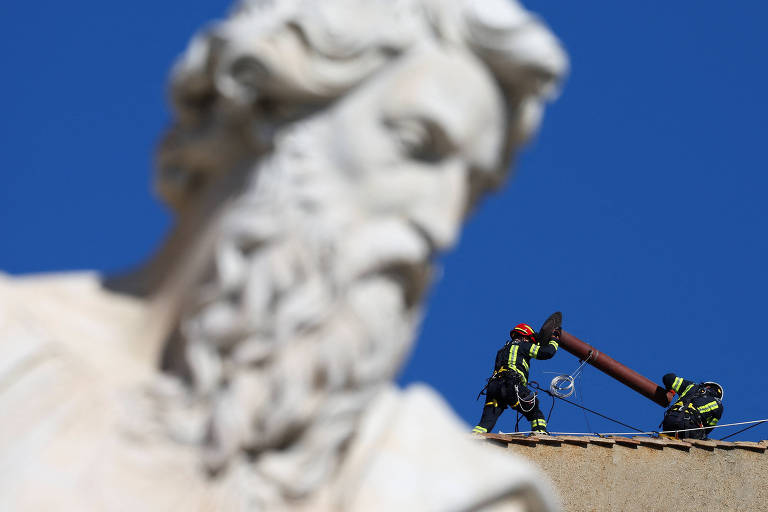 A imagem mostra uma escultura de um homem com barba em primeiro plano, enquanto ao fundo, dois bombeiros estão trabalhando no telhado de um edifício. Um dos bombeiros está segurando um tubo, enquanto o outro parece estar ajudando. O céu está claro e azul, proporcionando um contraste com a escultura e a cena de trabalho.