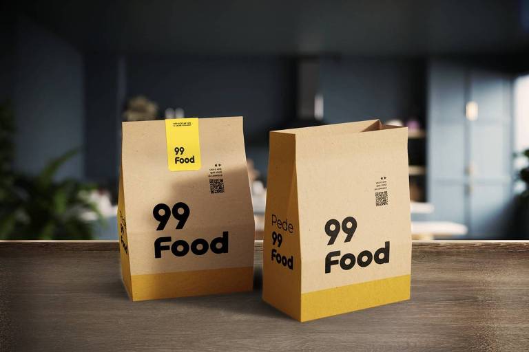 Duas embalagens de papel kraft, uma com uma etiqueta amarela e a outra sem, ambas com a inscrição '99 Food' em destaque. O fundo é desfocado, mostrando um ambiente interno com móveis e plantas.