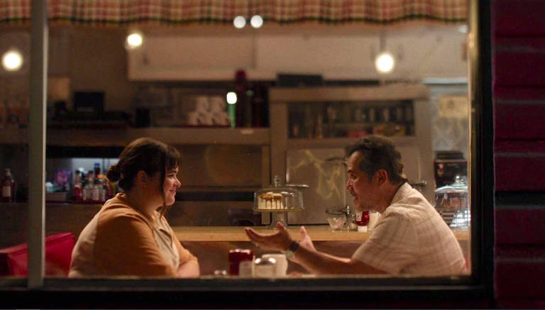 A imagem mostra duas pessoas sentadas em uma mesa de café, conversando. À esquerda, uma mulher com cabelo preso e um suéter claro sorri enquanto escuta. À direita, um homem com cabelo grisalho e camisa clara gesticula enquanto fala. Ao fundo, há uma vitrine com um bolo e prateleiras com garrafas e utensílios de café. A cena é iluminada suavemente, criando uma atmosfera acolhedora.