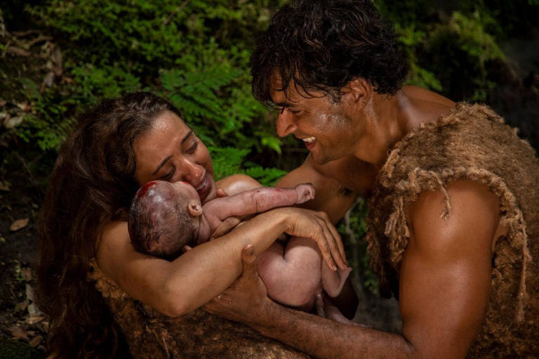 A imagem mostra um casal em um ambiente natural, cercado por vegetação. A mulher, com cabelo longo e solto, está segurando um bebê nos braços, enquanto expressa emoção. O homem, com cabelo escuro e curto, sorri para a mulher e o bebê, demonstrando felicidade. Ambos estão vestidos com peles, sugerindo um cenário pré-histórico. O fundo é composto por folhas verdes e uma atmosfera de natureza.