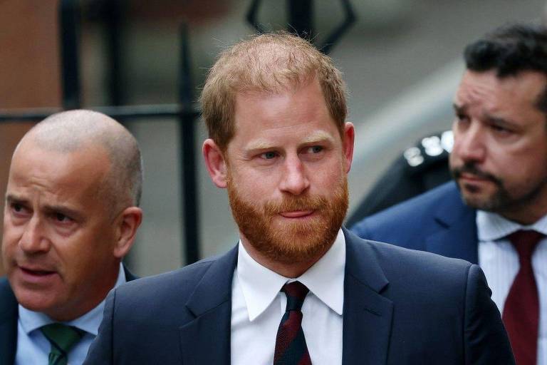 Um homem com cabelo curto e barba ruiva, vestido com um terno escuro e uma gravata listrada, está em primeiro plano. Ele parece sério e está cercado por dois homens, um deles com cabelo raspado e o outro com cabelo escuro. O fundo é desfocado, mas parece ser uma área urbana.