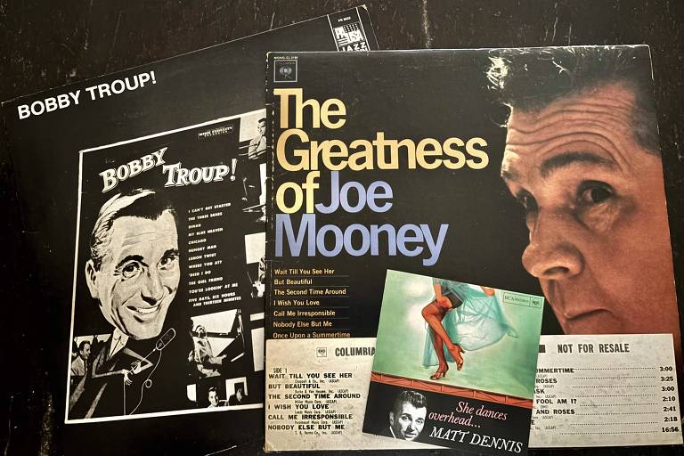 A imagem mostra três capas de discos de vinil sobre uma superfície escura. A capa superior é de 'Bobby Troup!', com uma foto em preto e branco de um homem sorridente. A capa do meio é de 'The Greatness of Joe Mooney', apresentando um close do rosto de um homem com cabelo grisalho. A capa inferior é de 'She Dances Overhead' de Matt Dennis, com uma imagem colorida de pernas femininas em uma saia azul. As capas contêm títulos e informações sobre as faixas dos discos.