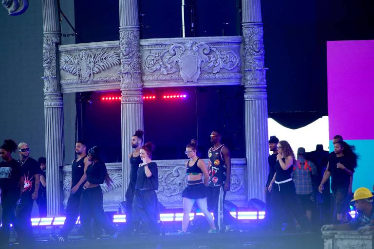 Um grupo de dançarinos e artistas está se apresentando em um palco decorado com colunas e luzes coloridas. Eles estão vestidos com roupas escuras e algumas pessoas usam acessórios como bonés. O fundo do palco tem uma tela iluminada com cores vibrantes, como roxo e azul. A atmosfera parece ser de um evento ao vivo, possivelmente um show ou festival.
