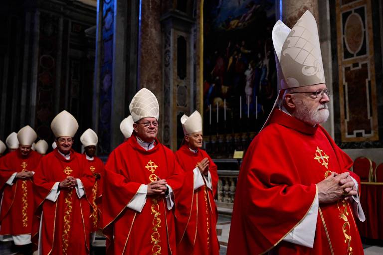 A imagem mostra um grupo de cardeais e bispos em vestes litúrgicas vermelhas, com mitras brancas, caminhando em um ambiente interno que parece ser uma catedral ou basílica. Eles estão em formação, com as mãos unidas em frente ao corpo, e há uma pintura religiosa ao fundo. A iluminação é suave, destacando os detalhes das vestes e do ambiente.