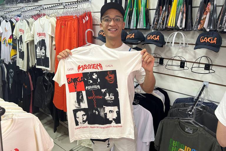 Um jovem está sorrindo enquanto segura uma camiseta branca com uma estampa colorida. A camiseta apresenta imagens em preto e vermelho, com o texto 'Madonna' e 'Gaga'. Ao fundo, há várias camisetas e bonés expostos em uma loja de roupas. O ambiente é bem iluminado e organizado, com uma variedade de roupas penduradas nas prateleiras.