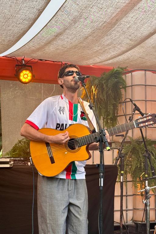 Um músico está se apresentando em um palco. Ele usa uma camisa branca com detalhes em verde e vermelho, e calças claras. O músico toca um violão e canta em um microfone. Ao fundo, há uma estrutura com plantas e uma iluminação suave. O ambiente parece ser ao ar livre, com uma cobertura de tecido.