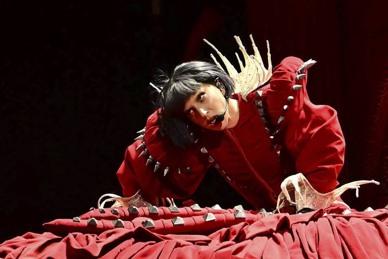 Uma artista de performance está em uma posição baixa, com o corpo apoiado em uma superfície vermelha. Ela usa um traje vermelho com detalhes pontiagudos e tem cabelo preto. A expressão facial é intensa, e a artista parece estar se movendo de forma dramática, com as mãos posicionadas de maneira expressiva. O fundo é escuro, destacando a figura da artista e o traje.