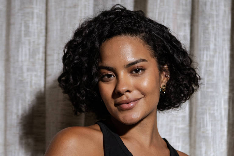 A imagem mostra uma mulher com cabelo cacheado e escuro, usando uma blusa preta. Ela está sorrindo levemente e posando em frente a um fundo de cortinas claras. A iluminação destaca seu rosto e a textura do cabelo.