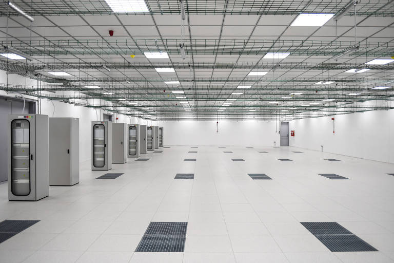A imagem mostra um ambiente amplo e vazio, com piso claro e teto elevado. Há várias unidades de armazenamento ou armários em um dos lados da sala, dispostos em linha. O teto é composto por uma grade metálica, e há iluminação fluorescente visível. O espaço parece ser uma sala de servidores ou um data center, com ventilação no chão.