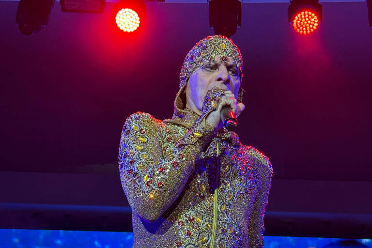Um artista está se apresentando em um palco iluminado com luzes vermelhas. Ele usa um traje brilhante e ornamentado, coberto de pedras preciosas e com um capuz. O artista segura um microfone e parece estar cantando, com uma expressão intensa no rosto.