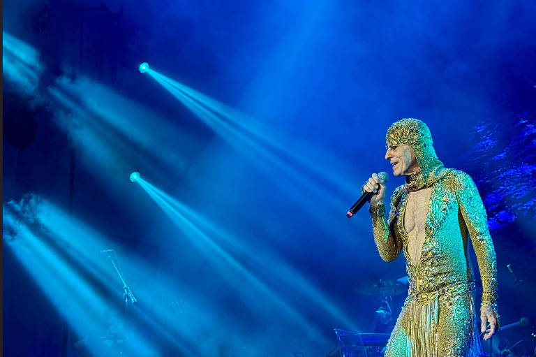 Um artista se apresenta em um palco iluminado com luzes azuis. Ele está vestido com um traje brilhante e dourado, que cobre todo o corpo, e segura um microfone enquanto canta. O fundo é desfocado, mas há uma iluminação dramática que destaca o artista.