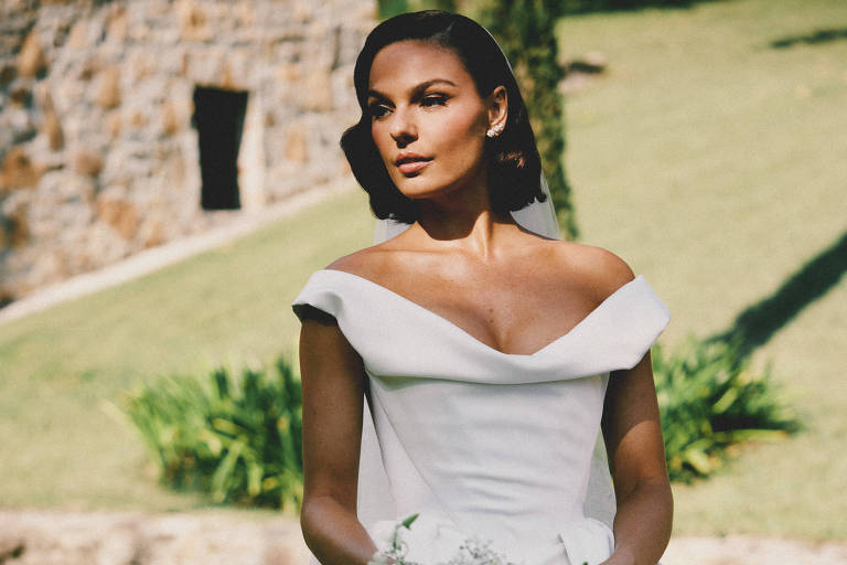 Uma mulher está em um ambiente ao ar livre, usando um vestido de noiva branco com um decote em forma de coração. Ela segura um buquê de flores brancas e tem um olhar sereno. O fundo é um gramado verde com uma construção de pedra ao fundo.