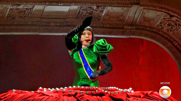 Uma pessoa está se apresentando em um palco, vestindo um traje verde com detalhes em azul e vermelho. O traje tem mangas longas e volumosas. A pessoa está em uma pose dramática, com uma mão levantada. O fundo do palco é decorado com um padrão arquitetônico e uma iluminação que destaca a figura. Há uma mesa com uma toalha vermelha na frente da pessoa.
