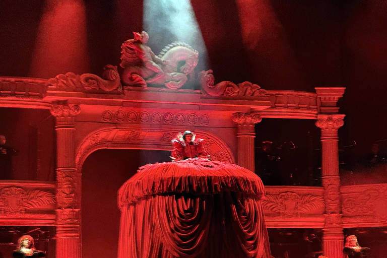 A imagem mostra uma cena de uma performance teatral com iluminação vermelha intensa. No centro, uma figura está sentada em uma grande estrutura coberta por um tecido vermelho, enquanto ao fundo há uma decoração elaborada com colunas e esculturas. A iluminação destaca a figura e cria um ambiente dramático, com outros performers visíveis nas laterais.