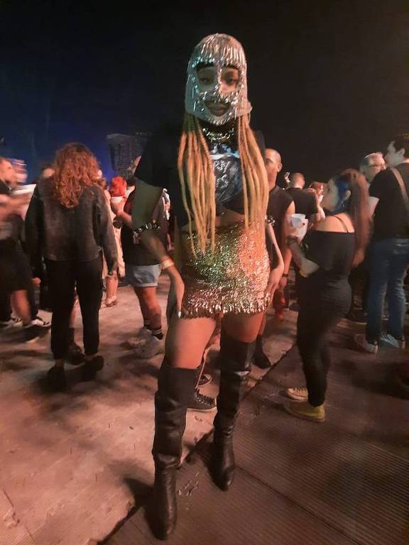 A imagem mostra uma pessoa em um evento noturno, vestindo um traje brilhante e uma máscara que cobre o rosto, com longos cabelos loiros. Ao fundo, há várias pessoas em um ambiente festivo, com luzes e uma atmosfera animada.