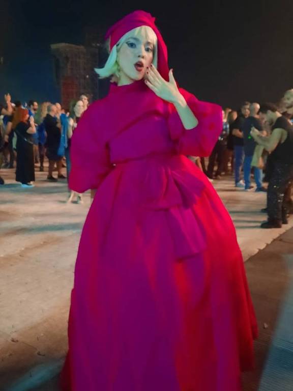 A imagem mostra uma mulher em um vestido longo e volumoso de cor rosa, com mangas bufantes e um cinto na cintura. Ela está usando um lenço rosa na cabeça e fazendo um gesto de beijo com a mão. Ao fundo, há várias pessoas em um ambiente noturno, com luzes e uma atmosfera festiva.