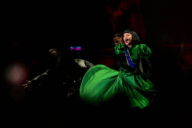 A imagem mostra uma cena de uma performance teatral, onde uma artista está vestida com um vestido verde vibrante. Ela está em movimento, com uma expressão intensa no rosto, enquanto um outro performer, vestido de preto, está ao fundo, aparentemente em uma pose dramática. O cenário é escuro, com iluminação que destaca os personagens.