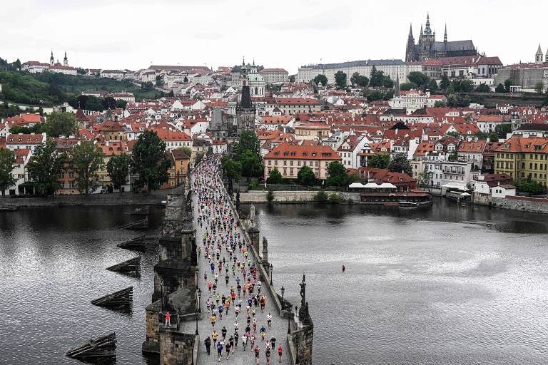 A imagem mostra a Ponte Carlos em Praga, cheia de pessoas caminhando. Ao fundo, há uma vista da cidade com prédios de telhados vermelhos e a famosa Catedral de São Vito. O céu está nublado e o rio Vltava reflete a paisagem ao redor.