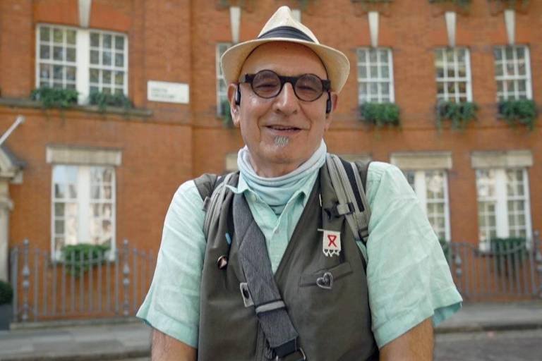 Een lachende man poseert voor een foto voor een rood bakstenen gebouw. Hij draagt een lichte hoed, een bril en een groen shirt met korte mouwen. De man draagt ook een vest met meerdere zakken en een sjaal om zijn nek. Op de achtergrond zijn grote ramen met potplanten te zien. Aan de voorkant van het gebouw bevindt zich een ijzeren hek.