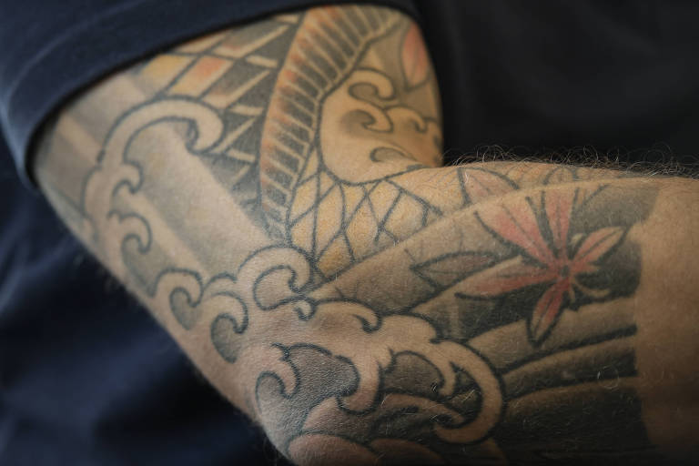 A imagem mostra um braço masculino com uma tatuagem detalhada que inclui ondas e uma flor. O braço está parcialmente coberto por uma camisa preta com um logotipo vermelho e azul, onde se lê 'Pacheco'.
