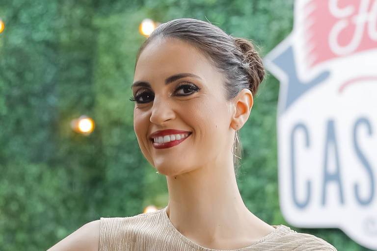 Uma mulher está posando em frente a um fundo verde com luzes brilhantes. Ela usa um vestido de cor clara e tem um sorriso no rosto. Ao fundo, há uma placa que diz 'Fábrica de Casamentos'.