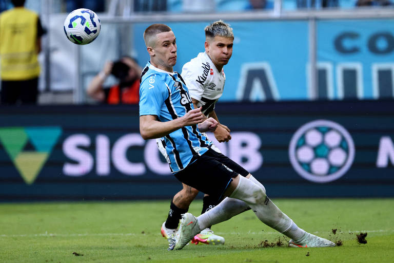 A imagem mostra um momento de um jogo de futebol, onde um jogador com camisa azul e preta está chutando a bola, enquanto um jogador com camisa branca observa. O campo está visivelmente molhado, e há um público ao fundo. O jogador da camisa azul parece estar em movimento, enquanto o jogador da camisa branca está em uma posição de espera.
