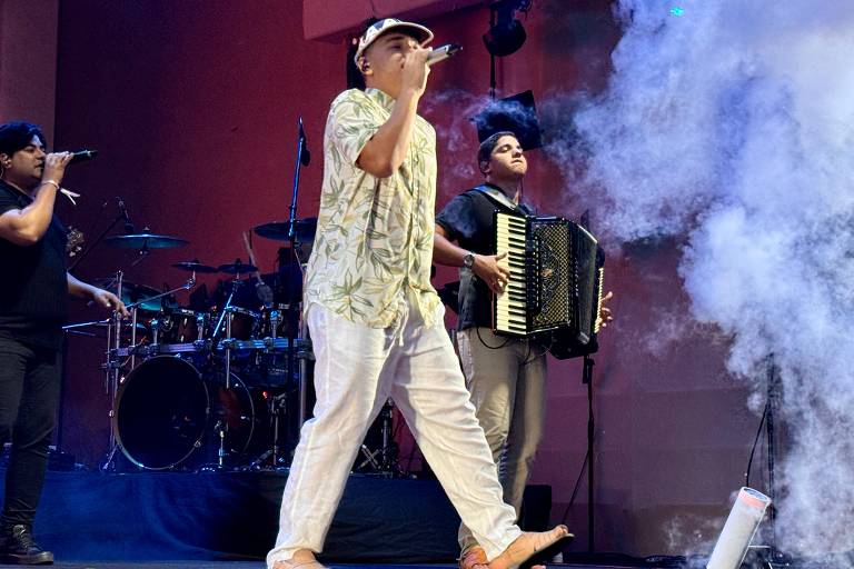 Um artista se apresenta no palco, cantando e segurando um microfone. Ele usa uma camisa estampada e calças brancas, além de um chapéu. Ao fundo, um músico toca acordeão e outro toca um instrumento de percussão. Há fumaça no palco e uma bateria visível ao fundo.