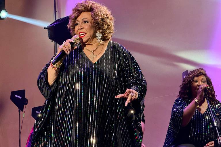 Uma cantora está se apresentando em um palco, usando um vestido preto brilhante com lantejoulas. Ela segura um microfone e sorri enquanto canta. Ao fundo, há uma outra cantora com um vestido semelhante. O cenário é iluminado com luzes de palco.