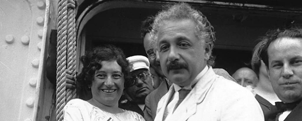 A imagem mostra um grupo de pessoas em um navio. No centro, um homem de cabelo grisalho e bigode, vestido com um paletó claro, está segurando um livro ou documento. Ao seu lado, uma mulher com cabelo cacheado e vestido claro sorri. Um menino à frente deles está sorrindo e fazendo um gesto com a mão. Ao fundo, outras pessoas estão visíveis, algumas parcialmente escondidas. O ambiente parece ser uma área de embarque ou desembarque.