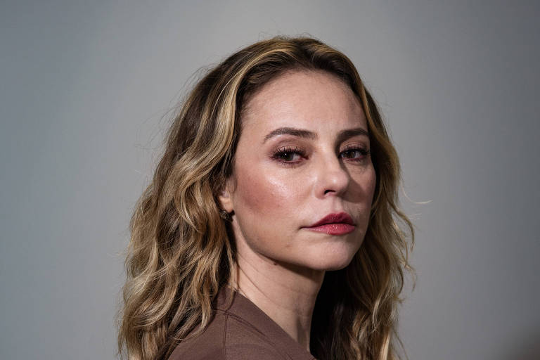 A imagem mostra uma mulher com cabelo ondulado e longo, usando uma blusa escura e um casaco marrom. Ela está posicionada de perfil, olhando para a frente, com uma expressão séria. O fundo é neutro, em tons de cinza.