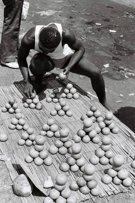 A imagem em preto e branco mostra um homem agachado em uma superfície coberta por uma esteira, organizando várias frutas redondas, possivelmente laranjas ou limões. O homem está usando uma camiseta sem mangas e parece concentrado em seu trabalho. O fundo é um espaço urbano, com sujeira visível no chão.