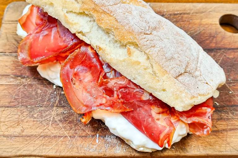 Um sanduíche montado em um pão ciabatta, recheado com fatias de presunto e tomate. O sanduíche está sobre uma tábua de madeira, com um fundo de madeira clara.