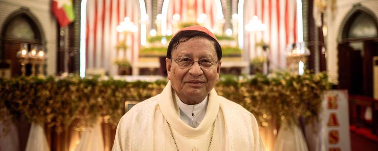 Um cardeal está posando em frente ao altar de uma igreja. Ele usa uma vestimenta religiosa branca com detalhes dourados e um cordão. Ao fundo, o altar está decorado com flores e velas, e há bandeiras visíveis nas laterais. A iluminação é suave, criando uma atmosfera solene.